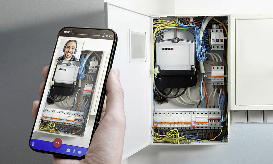 Eine Hand hält ein Smartphone mit dem e-mobilio Video-Installations-Check und zeigt einen Videoanruf mit einem Experten, um die Elektroinstallation und die sichtbare Verkabelung einer Wallbox zu besprechen, während sie auf einen offenen Schaltschrank zeigt.