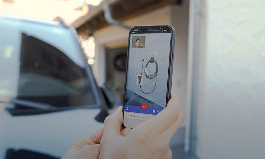 Eine Person bedient ein Smartphone mit dem Display des e-mobilio Video-Installations-Checks; auf dem Anrufbildschirm sind ein Headset und ein kleiner Video-Feed zu sehen, im Hintergrund suggeriert ein weißes Fahrzeug einen laufenden Installationsservice.