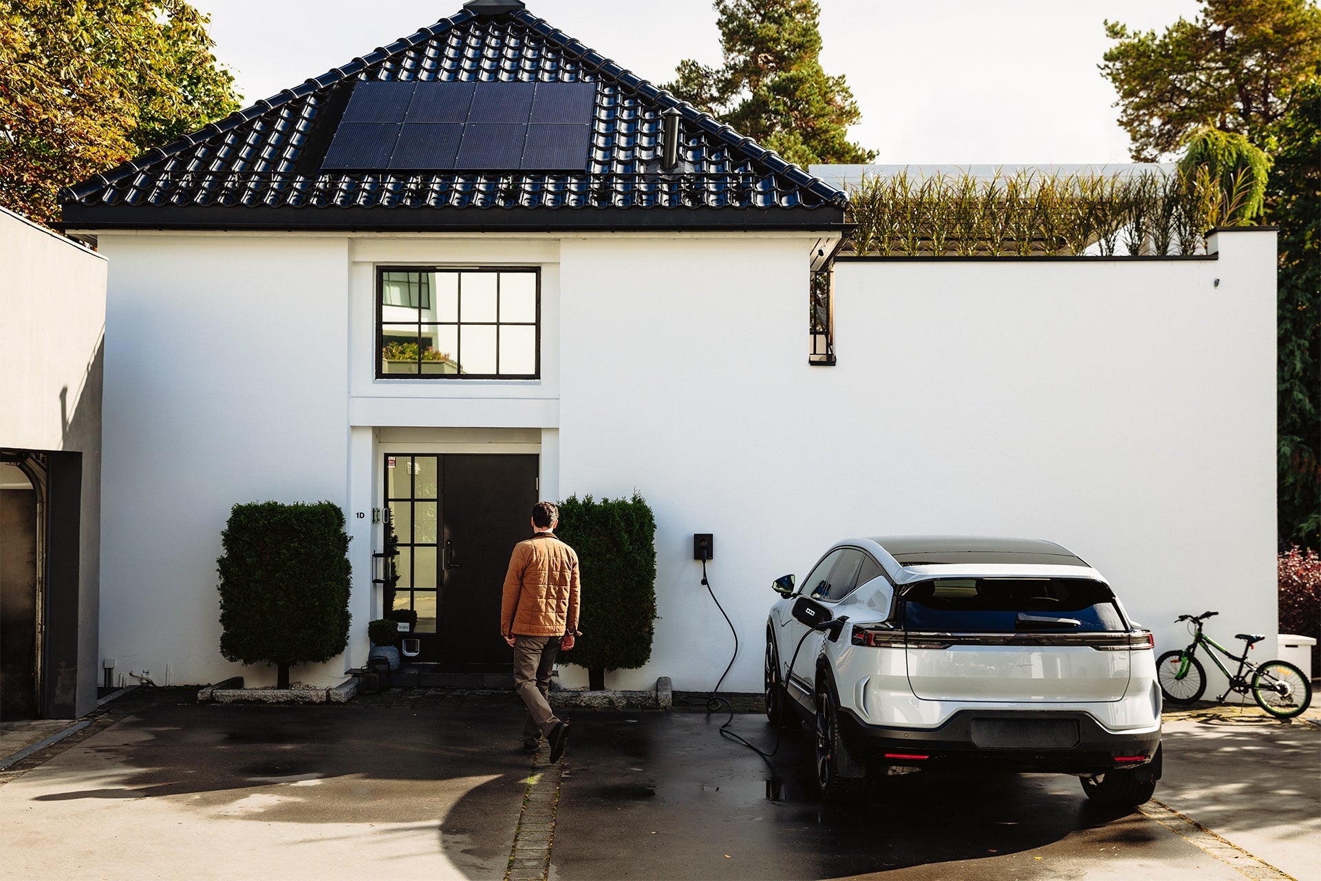 Ein Mann geht auf ein modernes weißes Haus mit Solarzellen zu, während in der Einfahrt ein Elektroauto an einem Zaptec Go 2 von Zaptec aufgeladen wird und ein grünes Fahrrad an der Wand steht.