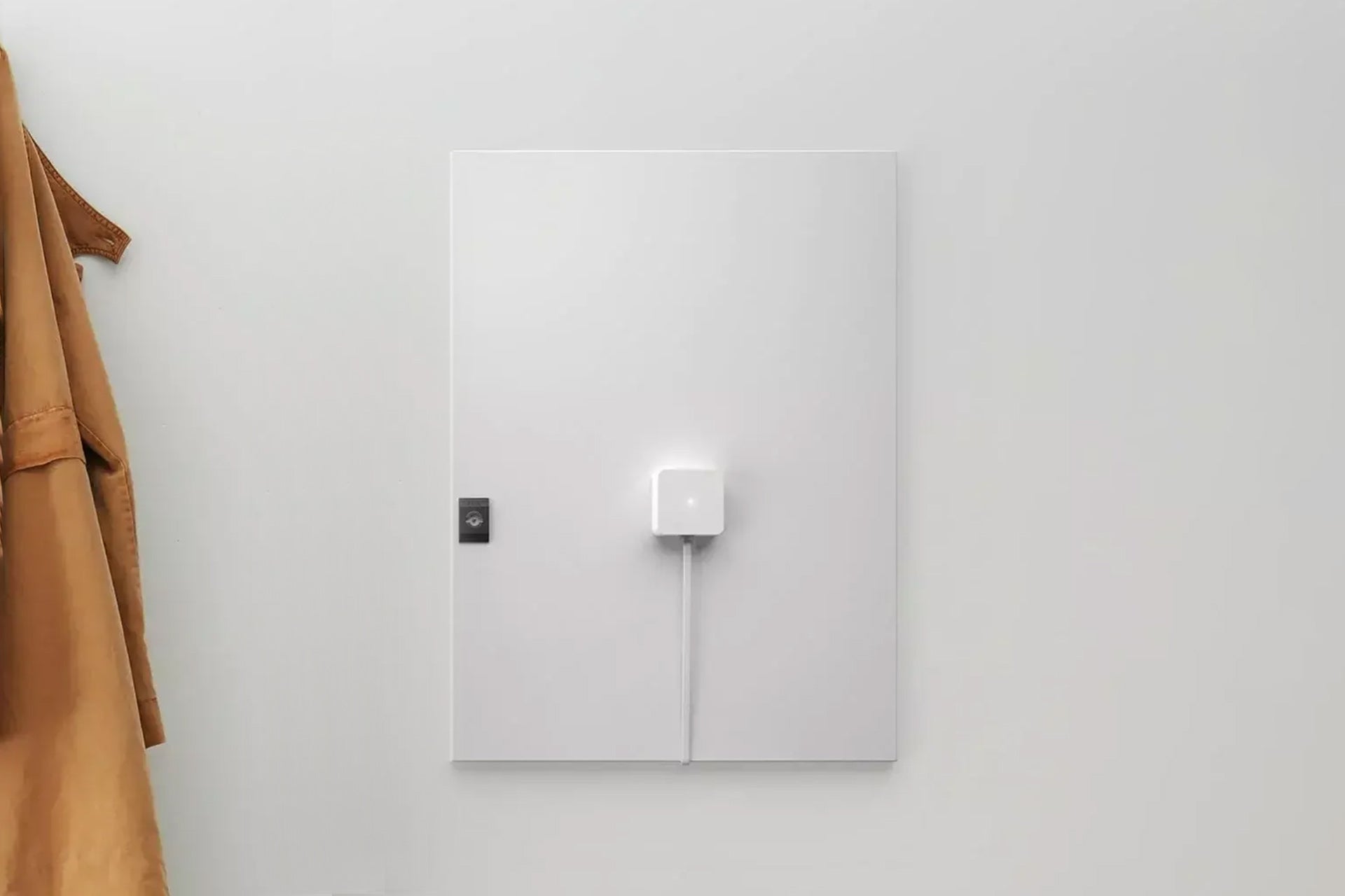 Ein weißes, quadratisches Wandpanel mit einem Zaptec Sense Gateway Kit von Zaptec und angeschlossenem Kabel ist an einer schlichten weißen Wand montiert – perfekt für dynamisches Lastmanagement beim Laden von Elektrofahrzeugen. Am linken Rand hängt ein brauner Mantel.