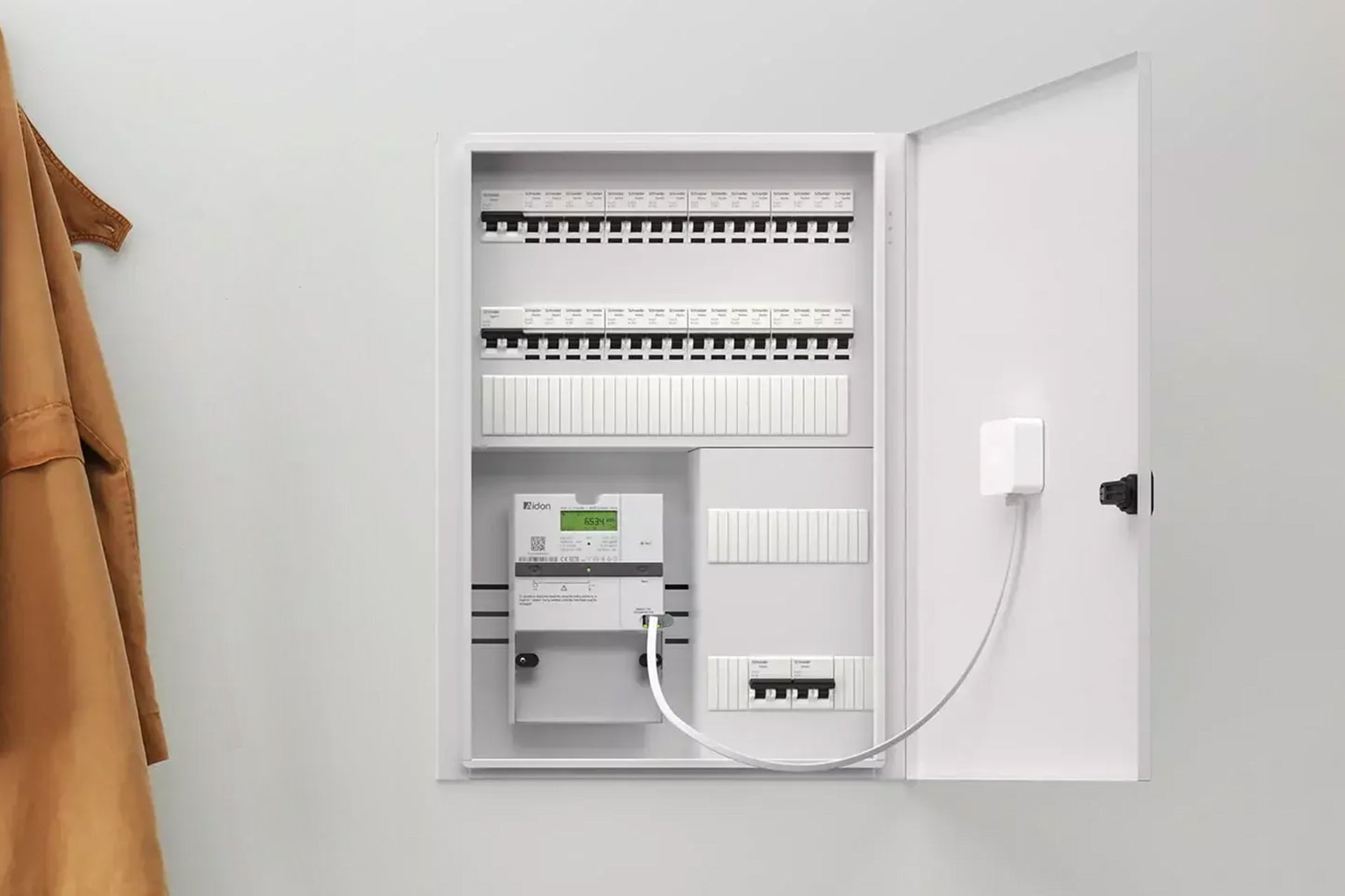 Ein offener Schaltschrank enthält Leistungsschalter, einen digitalen Zähler und das Zaptec Sense Gateway Kit von Zaptec für dynamisches Lastmanagement beim Laden von Elektrofahrzeugen. Links an der Wand hängt ein brauner Mantel.