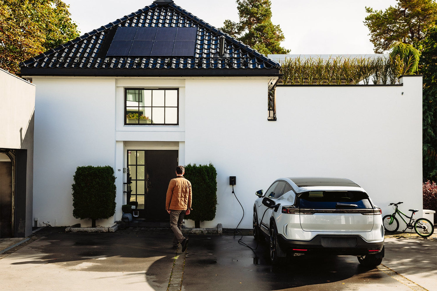 Ein Mann geht auf ein modernes weißes Haus mit Solarzellen zu, während in der Einfahrt ein Elektroauto an einem Zaptec Go 2 von Zaptec aufgeladen wird und ein grünes Fahrrad an der Wand steht.