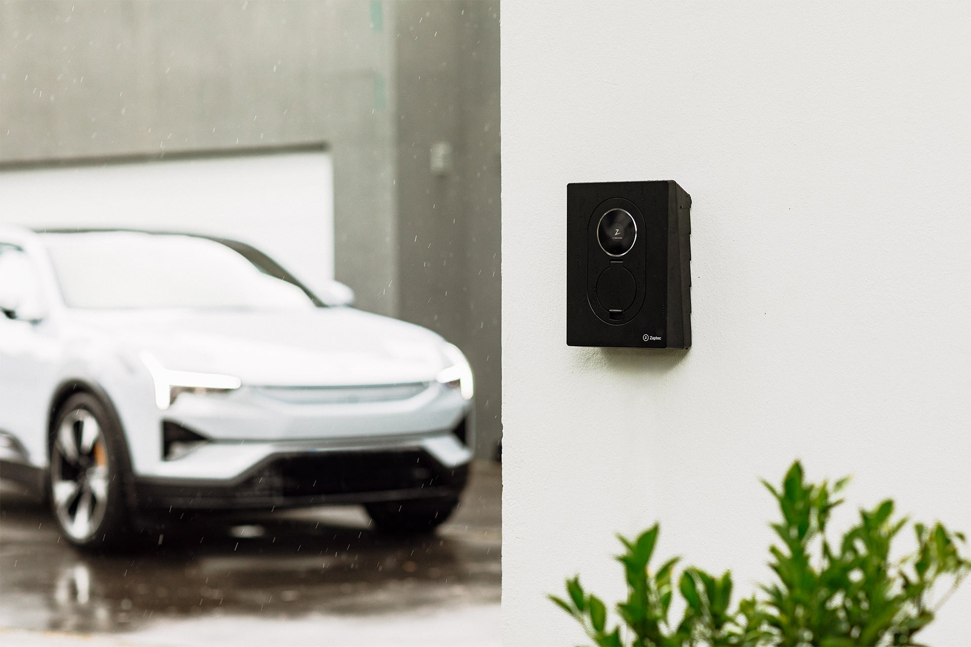 Ein schwarzer Zaptec Go 2 von Zaptec für bidirektionales Laden ist an einer weißen Wand montiert, im Hintergrund parkt an einem regnerischen Tag ein weißes Auto.