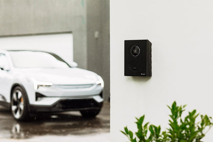 Ein schwarzer Zaptec Go 2 von Zaptec für bidirektionales Laden ist an einer weißen Wand montiert, im Hintergrund parkt an einem regnerischen Tag ein weißes Auto.