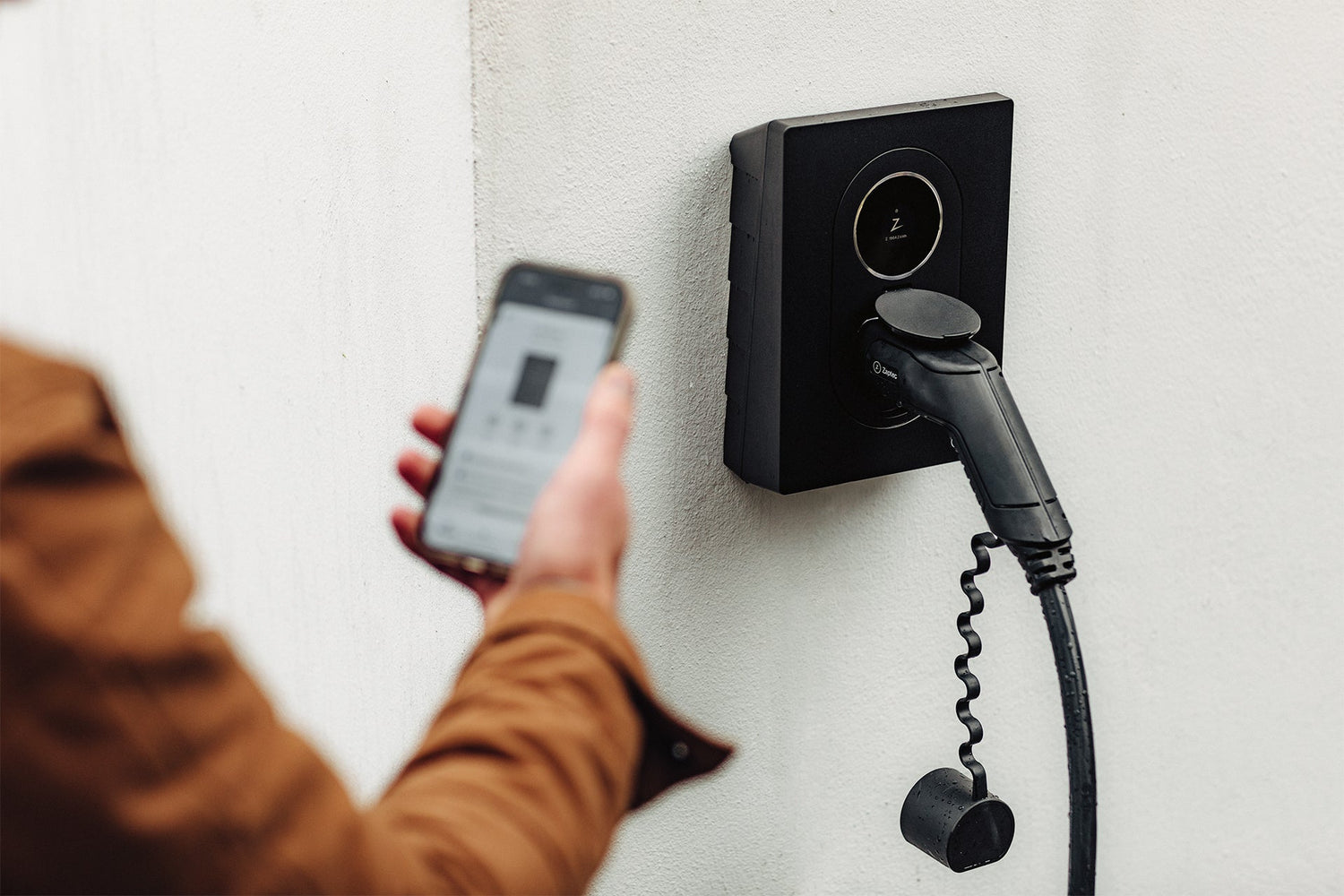 Eine Person schließt ein Ladegerät für ein Elektrofahrzeug an, während sie ein Smartphone hält. Dabei wird der Zaptec Go 2 von Zaptec mit bidirektionalem Laden hervorgehoben, der an einer weißen Wand montiert ist.