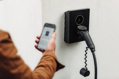 Eine Person schließt ein Ladegerät für ein Elektrofahrzeug an, während sie ein Smartphone hält. Dabei wird der Zaptec Go 2 von Zaptec mit bidirektionalem Laden hervorgehoben, der an einer weißen Wand montiert ist.